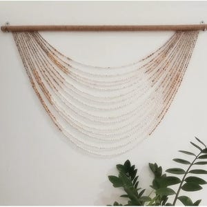 Op de afbeelding: Een decoratieve wanddecoratie met strengen kleine, witte en bruine kralen die aan een houten staaf zijn opgehangen. De kralen zijn gerangschikt in een gebogen, trapsgewijs patroon, waardoor een gestructureerd, vloeiend ontwerp ontstaat. De wanddecoratie is aan een witte muur bevestigd.