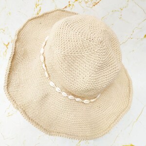 May include: A beige, woven sun hat with a wide brim. The hat is decorated with a string of small, white seashells around the crown. The hat is designed for sun protection and beachwear.