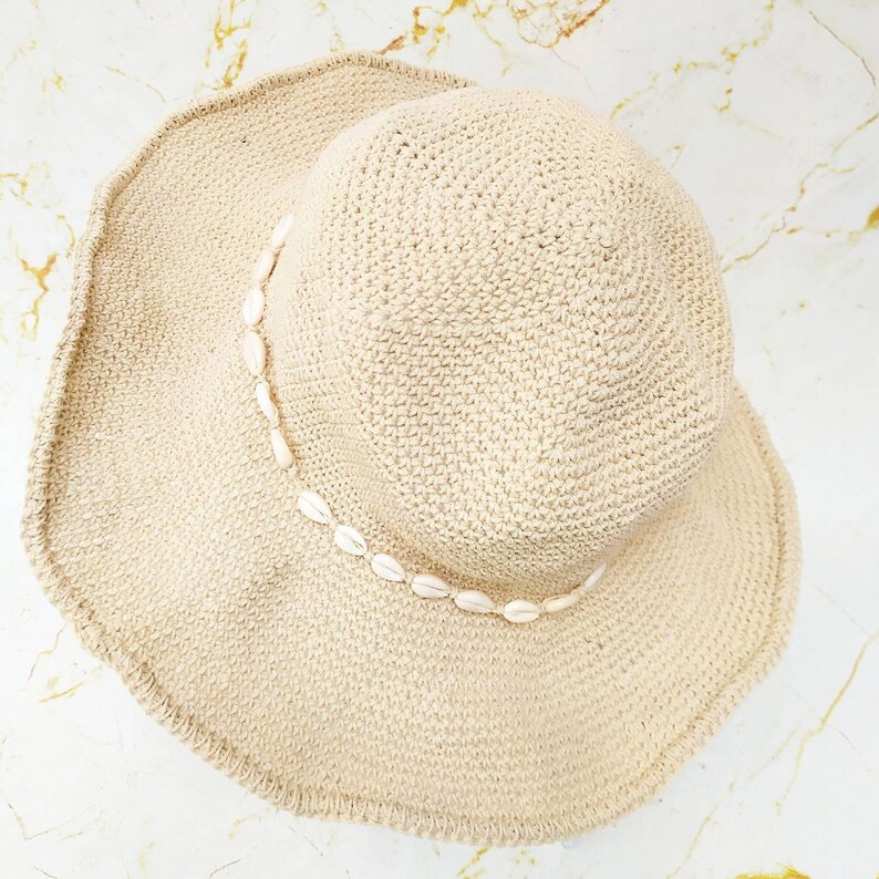 May include: A beige, woven sun hat with a wide brim. The hat is decorated with a string of small, white seashells around the crown. The hat is designed for sun protection and beachwear.