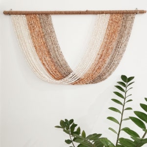 Op de afbeelding: Een kralen wanddecoratie, opgehangen aan een natuurlijke houten stok met jute touw. De kralenstrengen creëren een gestreept patroon in wit, beige en bruin. De wandkunst hangt aan een witte muur, met groene planten op de voorgrond.