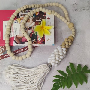 Puede incluir: Un borla blanca cuelga de una guirnalda de cuentas de madera con un racimo de conchas blancas. La guirnalda está drapeada sobre un libro con una flor amarilla en la parte superior.