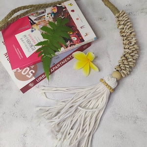 Può includere: Una collana fatta a mano con un'estetica naturale e bohémien. La collana presenta un cordoncino di iuta intrecciata, una fila di piccole conchiglie, perline di legno e un nappa di cotone bianco. Una felce e un fiore giallo sono posti vicino alla collana.