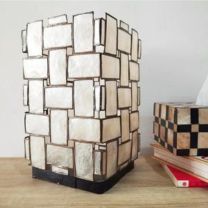 Può includere: Un paralume rettangolare in madreperla intrecciata bianco e oro. Il paralume poggia su una base nera.