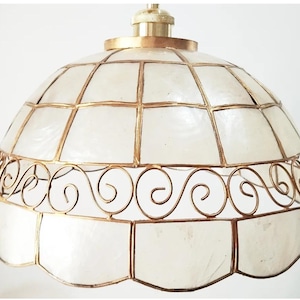 Könnte beinhalten: Eine weiße und goldene Pendelleuchte mit geometrischem Design. Die Leuchte besteht aus einem weißen Material mit einem goldenen Metallrahmen. Die Leuchte hat einen gewellten Rand.