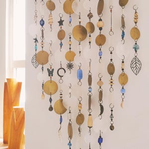 May include: A wall hanging featuring a horizontal wooden branch suspending multiple vertical strands of mixed materials. The strands include circular brass discs, translucent white shell-like discs, blue glass beads, and metal charms shaped like leaves, stars, and butterflies.