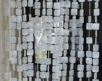 May include: A white beaded curtain made of square-shaped shells. The curtain is hanging in a doorway and is made of multiple strands of beads.