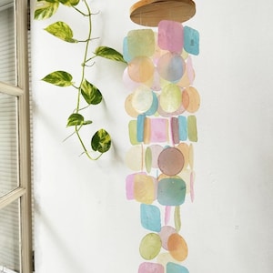 Könnte beinhalten: Ein farbenfroher Windspiel mit einem Holzaufsatz und Strängen aus schillernden, pastellfarbenen Muschelteilen in verschiedenen Formen. Das Windspiel hängt vor einer weißen Wand, mit einer grünen Rankpflanze im Hintergrund.
