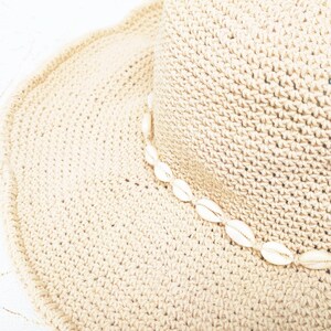 May include: A beige woven sun hat with a wide brim. The hat is decorated with a string of white seashells around the crown. The hat is made of a textured, crocheted material.