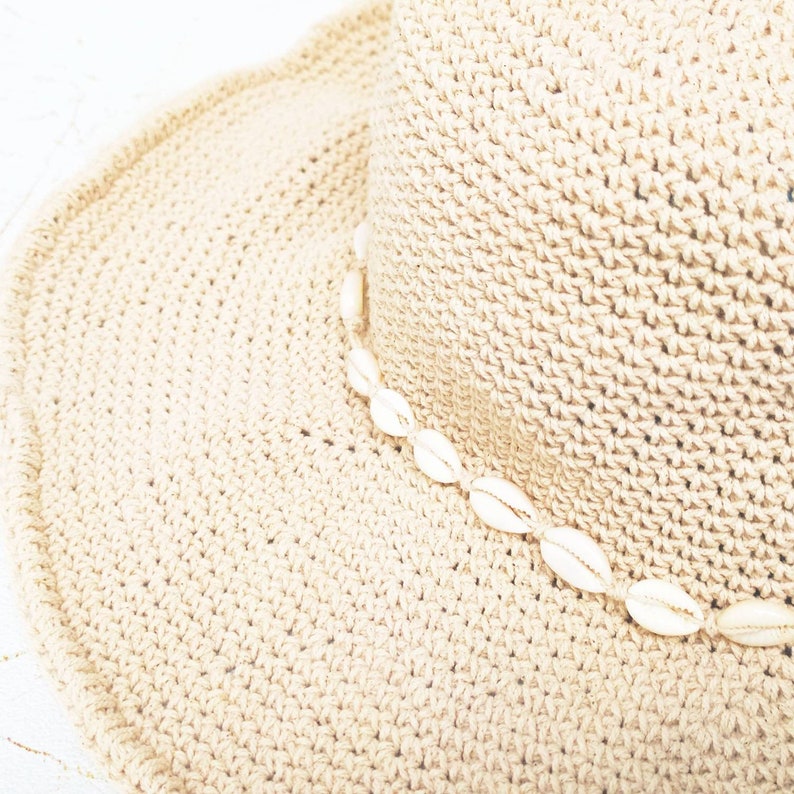 May include: A beige woven sun hat with a wide brim. The hat is decorated with a string of white seashells around the crown. The hat is made of a textured, crocheted material.