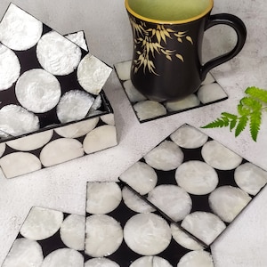 Könnte beinhalten: Satz mit sechs schwarzen und weißen Untersetzern mit einem kreisförmigen Muster. Die Untersetzer sind aus Perlmutt gefertigt und haben eine glänzende Oberfläche. Eine schwarze Tasse mit einem Blattgold-Design steht auf einem der Untersetzer.