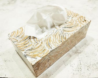 May include: A rectangular tissue box with a carved design. The box is made of light-coloured wood with a white and gold pattern. White tissues are visible, pulled from the top opening. The box is on a white surface.