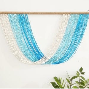 Op de afbeelding: Een wanddecoratie gemaakt van witte en blauwe kralen, gerangschikt in een verloop. De kralen zijn geregen aan een koord van natuurlijke vezels en opgehangen aan een houten lat.