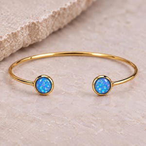 May include: A gold-toned open cuff bracelet featuring two circular settings at each end. Each setting contains a round, vibrant blue opal-like stone with iridescent flecks of light, creating a bright and reflective appearance.
