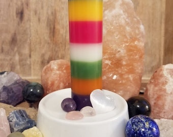 May include: A multi-colored candle with layers of blue, yellow, red, white, green, orange, and purple wax. The candle is sitting on a white pedestal with various crystals and stones around it.
