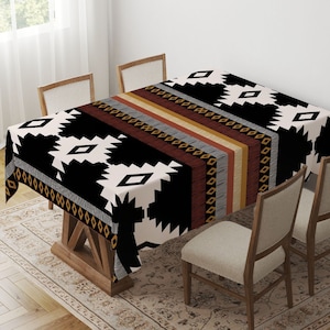 Op de afbeelding: Een rechthoekig tafelkleed met een geometrisch patroon in zwart, wit, bruin, grijs en geel. Het tafelkleed ligt op een houten tafel, omringd door vier stoelen. Het patroon bevat ruit- en zigzagvormen.