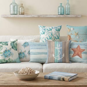 May include: A collection of nine square throw cushions arranged on a sofa, featuring various coastal and nautical designs including sea turtles, anchors, starfish, and the text "BEACH LOVE" in shades of blue, white, tan, and green.