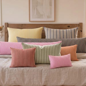 May include: A collection of rectangular and square throw pillows featuring vertical stripes in various colours including yellow, blue, black, pink, green, and orange, arranged on a bed with a light-coloured linen duvet and a wooden headboard.