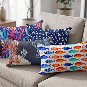 May include: Five decorative throw pillows with nautical themes rest on a gray sofa. Designs include coral, anchors, fish, and marine life in shades of blue, orange, and white. The pillows vary in shape and size, adding visual interest to the scene.