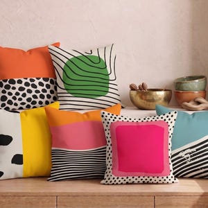 Puede incluir: Una colección de seis cojines cuadrados con diversos patrones y colores, incluyendo naranja, amarillo, rosa, azul turquesa, lunares blancos y negros, y rayas horizontales negras, dispuestos sobre una superficie de madera.
