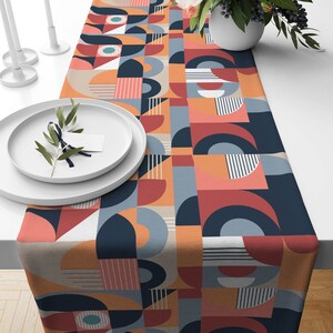 Könnte beinhalten: Ein geometrisch gemusterter Tischläufer mit einer Mischung aus orange, rot, blau und weißen Farben. Der Läufer ist aus gewebtem Stoff gefertigt und hat ein modernes Design.