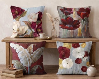 May include: Four decorative throw pillows with floral designs. One pillow features white and burgundy magnolia blossoms on a light blue background. Another displays red and white flowers. The other two pillows have red and white flowers and large leaves.