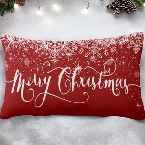Puede incluir: Un cojín rectangular rojo con copos de nieve blancos y la frase "Merry Christmas" en cursiva. Una decoración festiva para las fiestas navideñas.