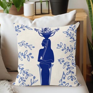 Puede incluir: Coj&iacute;n decorativo con una silueta azul de una persona que lleva un cuenco de flores. El fondo crema est&aacute; adornado con motivos florales azules. Un elemento decorativo para el sal&oacute;n o el dormitorio.