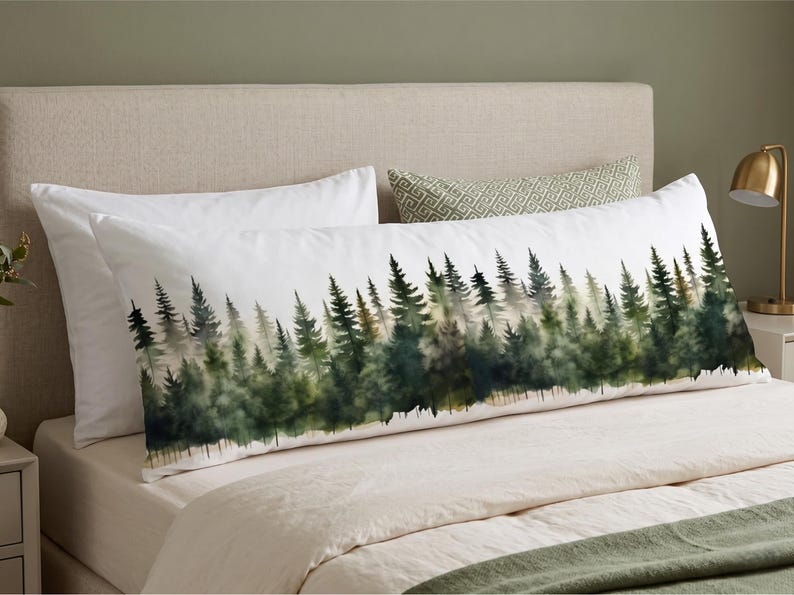 May include: A white body pillow with a watercolor forest scene. The design features a line of green and dark green trees against a white background. The pillow is on a bed with white sheets and a green blanket.