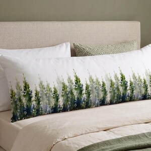 May include: A long, white body pillow with a watercolor design of green and blue evergreen trees. The pillow is on a bed with white sheets and a beige headboard. A smaller patterned pillow is behind it.