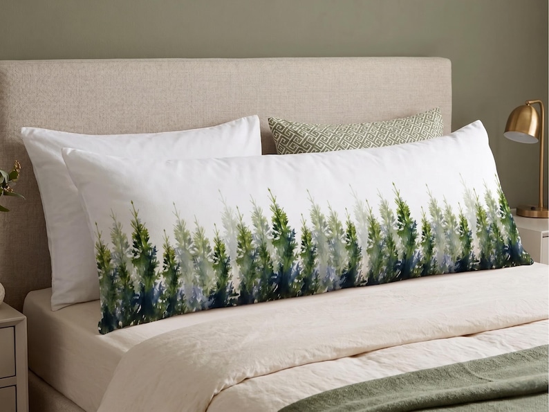 May include: A long, white body pillow with a watercolor design of green and blue evergreen trees. The pillow is on a bed with white sheets and a beige headboard. A smaller patterned pillow is behind it.