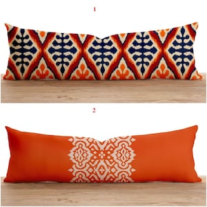 Pode incluir: Duas almofadas decorativas longas e retangulares. A almofada superior tem um padrão geométrico em azul marinho, laranja e creme. A almofada inferior é laranja lisa com um desenho branco. Ambas as almofadas estão sobre uma superfície de madeira.