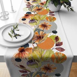May include: A white table runner with a floral design. The runner features watercolor-style flowers in shades of orange and brown, with green leaves and abstract shapes. The design is printed on a white background.