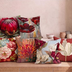 May include: A collection of six square throw pillows featuring vibrant floral prints. The designs include a large pink protea, deep red poppies, a bouquet of pink and blue peonies, an orange pincushion flower, white magnolias, and a single white tulip.