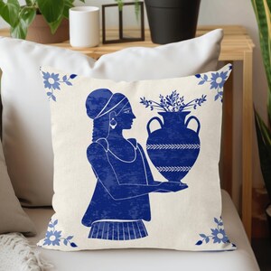Puede incluir: Coj&iacute;n decorativo con un dise&ntilde;o azul y blanco. La imagen presenta una ilustraci&oacute;n azul de una mujer sosteniendo un jarr&oacute;n lleno de vegetaci&oacute;n. Acentos florales enmarcan los bordes del coj&iacute;n, que est&aacute; sobre una superficie blanca.