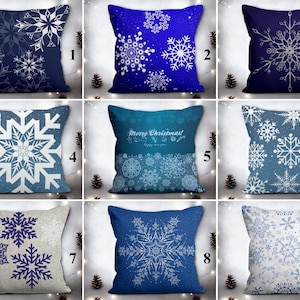 Könnte beinhalten: Neun dekorative Kissen mit Schneeflockenmustern in verschiedenen Blau- und Weißtönen. Jedes Kissen hat ein anderes Schneeflockenmuster, einige mit Text. Die Kissen sind auf einem hellen Hintergrund mit Tannenzapfen angeordnet.