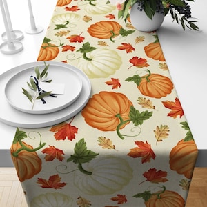 May include: A beige table runner featuring a repeating pattern of orange and white pumpkins, along with red, orange, and yellow autumn leaves. Green vines and leaves accent the pumpkins. The runner is placed on a white table, with white plates and candles visible.