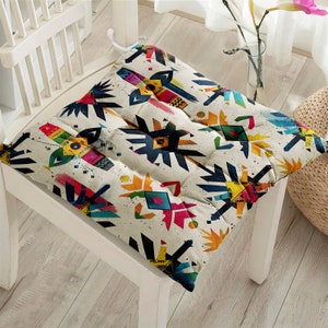 May include: Square chair cushion with a cream background and a colorful geometric pattern. The design features shapes in shades of blue, yellow, pink, and green. The cushion is quilted and sits on a white wooden chair.