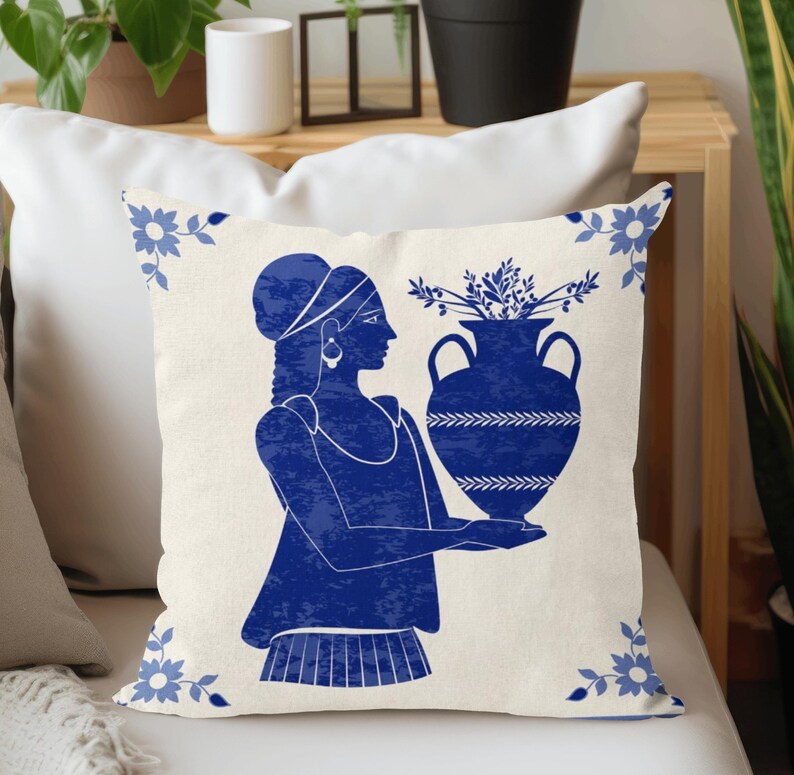 Puede incluir: Coj&iacute;n decorativo con una ilustraci&oacute;n azul y blanca. Muestra a una mujer sosteniendo un jarr&oacute;n lleno de follaje. Dise&ntilde;os florales adornan las esquinas. El coj&iacute;n est&aacute; sobre una superficie blanca.