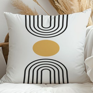 Puede incluir: Cojín cuadrado blanco con un diseño geométrico. El diseño presenta líneas arqueadas negras y una forma ovalada dorada sobre un fondo blanco. El cojín está sobre una silla de madera con tela blanca.