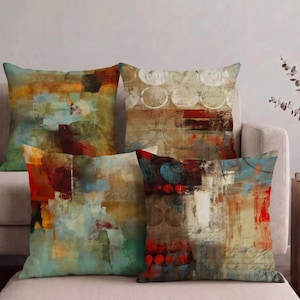 Puede incluir: Cuatro cojines cuadrados con diseños abstractos, texturas en capas y colores que incluyen tonos azul verdoso, naranja quemado, rojo intenso, marrón y crema. Los patrones incorporan círculos geométricos, efectos de pinceladas y marcas de escritura.