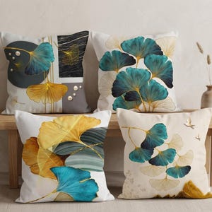 Può includere: Quattro cuscini quadrati con motivi a foglie di ginkgo in tonalità verde acqua, oro e nero. I design includono forme geometriche, texture marmorizzate e piccoli accenti simili a perle su uno sfondo color crema, disposti in una griglia.