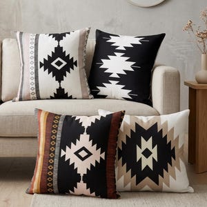 Puede incluir: Un conjunto de cuatro cojines cuadrados con patrones geométricos en negro, blanco y beige. Los diseños incluyen formas de diamante, rayas verticales y líneas en zigzag. Los cojines están dispuestos en grupo, mostrando una variedad de combinaciones de colores contrastantes y motivos simétricos en sus fundas de tela.