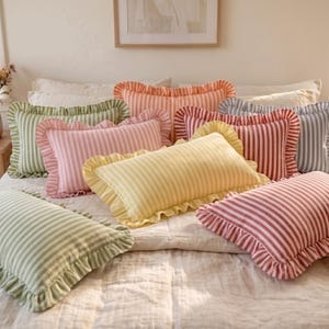 Può includere: Una collezione di cuscini rettangolari caratterizzati da righe verticali in vari colori pastello, tra cui giallo, rosa, verde e rosso. Ogni cuscino è rifinito con un bordo decorativo in tessuto arricciato lungo i lati, creando una texture morbida.