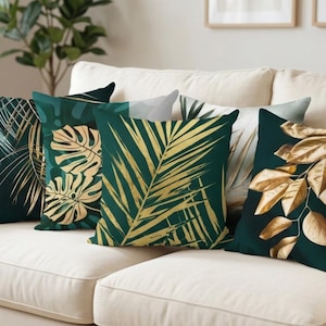 Op de afbeelding: Vijf decoratieve kussens met botanische ontwerpen liggen op een crèmekleurige bank. De kussens hebben donkergroene achtergronden met gouden en witte bladerpatronen. De ontwerpen omvatten palmbladeren en monstera-bladeren, wat een tropisch tintje geeft.