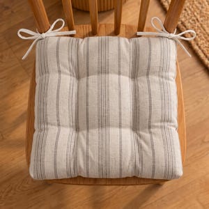 May include: A square seat cushion featuring a light beige fabric with vertical dark gray pinstripes. The cushion is tufted with nine distinct sections and includes white fabric ties at the two back corners to secure it to a wooden chair.