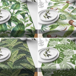 May include: Four different table runners with tropical leaf patterns. The runners are white with green leaves and feature different designs. The first runner has large green leaves, the second has smaller leaves and coconuts, the third has a dark green background with large leaves, and the fourth has large green monstera leaves.