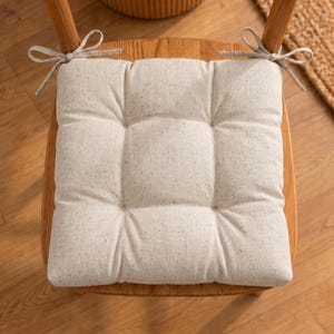 Puede incluir: Un cojín de asiento cuadrado de color blanco roto con una textura sutilmente moteada, que presenta cuatro hendiduras acolchadas y dos lazos de tela en las esquinas superiores para sujetarlo a una silla de madera. Es de tela tejida.