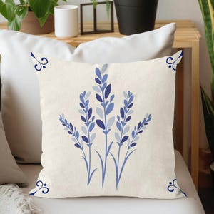 Puede incluir: Coj&iacute;n cuadrado color crema con un dise&ntilde;o floral azul. El dise&ntilde;o incluye ramitas de lavanda azules estilizadas y detalles decorativos en las esquinas. El coj&iacute;n est&aacute; colocado sobre una superficie blanca.