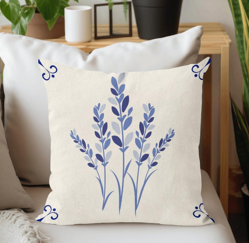 Puede incluir: Coj&iacute;n cuadrado color crema con un dise&ntilde;o floral azul. El dise&ntilde;o incluye ramitas de lavanda azules estilizadas y detalles decorativos en las esquinas. El coj&iacute;n est&aacute; colocado sobre una superficie blanca.