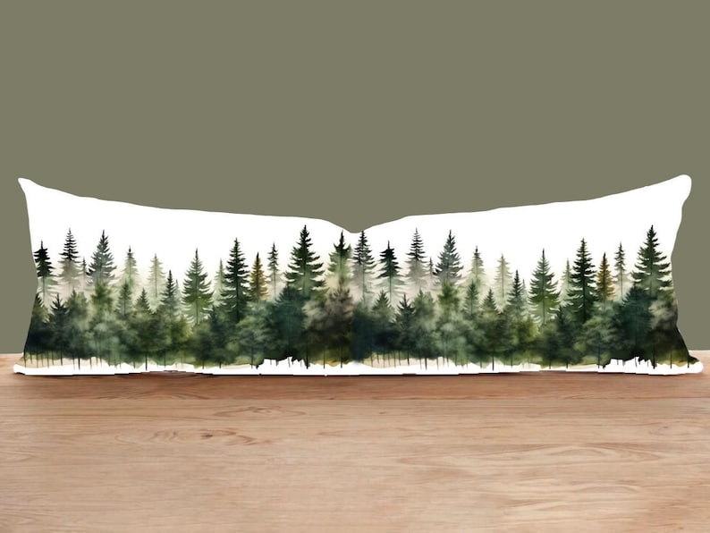 May include: A rectangular decorative pillow featuring a watercolor painting of a forest scene. The artwork depicts a line of evergreen trees in varying shades of green, with a misty background. The pillow is on a wooden surface.